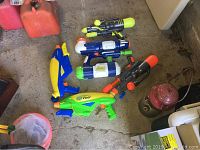 Five plastic water guns on a concrete floor including yellow, green, blue, white and black colors.