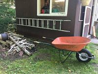 Photo of the metal wheelbarrow and aluminum extension ladder against a brown building wall with some logs on the grass.