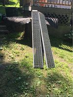 Pair of metal car ramps positioned outdoors on grass with diamond perforated grip surfaces.