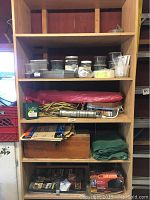 Four shelves with multiple containers of screws and nails, yellow coiled extension cord, wooden toolbox with tools, green folded fabric likely solar panel cover, and packaged items on bottom shelf.