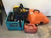 Crate containing chaps, small oil jug, hatchet, tarp, fuel canister, face shield and ear muffs alongside orange chainsaw case