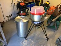 Wide view showing stainless steel deep fryer pot, three-legged burner stand, batter mixing bowl with cover, and additional pots in a workshop environment.