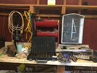 Overview of workstation bench showing metal case of drill bits, gloves, vises, red metal shelf with hardware, measuring ruler, and other assorted tools and items.