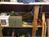 Shelf with white bucket holding plumbing/electrical hardware, green toolbox, yellow extension cord, gloves, red bench vise, saws, and level.