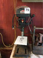 Front view of the Jobmate drill press showing the power switch, column, and adjustable table with user manual.