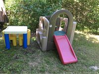 Outdoor kids play set including a tan Little Tikes slide with a red slide and green steps beside a small white table with blue legs and yellow chairs on grass.