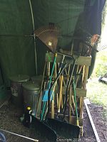 Photo showing three steel 20-gallon trash cans with lids stacked together near yard tools and a wooden stand.