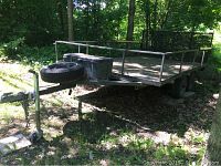 Front three-quarter view of trailer showing deck, railing, spare tire on tongue and lockable resin storage bin