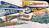 Photo of eight vintage USS naval ship pennants laid out on a white surface, showing colorful designs and ship illustrations.