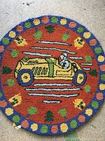 Full view of the round hooked rug showing the yellow vintage car with driver and decorative border of dots and leaves on an orange background.
