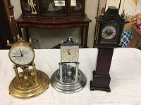 Three clocks displayed on a white cloth with wooden furniture background. Gold-tone and silver-tone anniversary clocks with pendulums on the left and center, tall dark grandfather clock replica on the right.