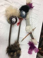 Three Indigenous decorative pieces on a white fabric surface including two ceremonial sticks with fur and circular emblems, and a small bow with two arrows decorated with feathers and fur.