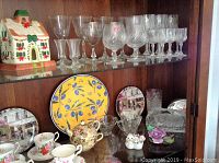 Wide shot of glassware and decorative ceramics in a wooden display cabinet; includes clear crystal glasses of various sizes, teacups, saucers, and a decorative yellow plate with blue olive motif.