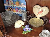 Photo showing glass serving platters, ceramic animal sculptures, pewter cup, heart-shaped Welcome plaque, and other decorative items on wooden shelf.