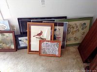 Nine framed artworks arranged in a cluster on the floor showing a variety of prints and one paint by numbers piece with text and floral decoration.