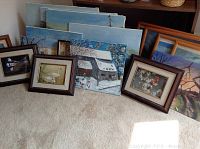 Collection of 16 artwork pieces leaning against a wall, largest is a winter house scene, includes four wooden framed bird prints and several other paintings mostly of natural and rural scenes.