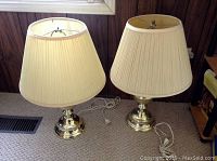 Pair of two brass base table lamps with beige pleated shades standing side by side on carpeted floor.