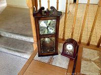 Two quartz table clocks placed on a table near staircase. The taller clock has a wood base and decorative carved top with missing trim. The smaller clock is rounded wood with glass front.