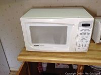 Front and side view of white Kenmore microwave oven with control panel, placed on wooden countertop against wallpapered wall.