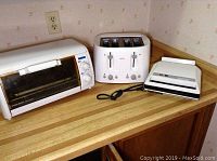 Three small kitchen appliances on a wooden countertop under a power outlet: Black and Decker toaster oven on left, Sunbeam four-slice toaster in center, Proctor Silex sandwich toaster on right.