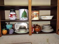 Photo showing kitchen shelves with Christmas ice bucket, snowman tin, assorted dishes, brown teapot, and small bowls