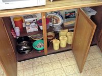 Wooden cabinet open showing vintage Tupperware canisters, lettuce crisper without lid, assorted pots and pans, salad spinner and other kitchen items