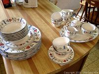 Display of the majority of the china set including stacked plates, bowls, tea cups, saucers, and serving pieces on a wooden table.