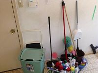 Photo showing green trash can, mops, step stool, and cleaning supplies arranged on floor against wall.