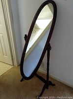 Full view of the oval wooden floor mirror standing on a carpeted floor, showing its frame, stand, and base.