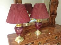 Two lamps shown side by side on a wooden dresser surface, with the lights off and cords visible, reflecting in a mirror behind.