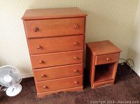 Wood dresser next to night table on carpeted floor with fan next to them.
