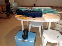 Photo shows white plastic outdoor table with two stools, vintage blue Coleman cooler below table, camp chair bag, rolled umbrella, camp pillow and packaged rain pants on top of table