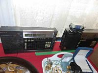 Two Sony ICF-2010 radios side by side on red surface, one fitted with AC adapter cord.