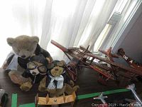 Group shot of three aviation-themed stuffed bears in pilot outfits with goggles, scarves, and jackets, positioned near three wooden model airplanes on a wooden surface.