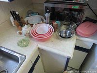 Overview of kitchen counter showing pink Pyrex mixing bowl set nested, stainless steel bowls, microwave bowl with pink lid, and butcher block knife set.