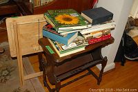 Wooden table with stack of assorted books including cooking and gardening titles, light wooden magazine rack visible behind.