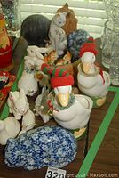 Photo showing two large white geese figurines with knit hats, rabbits, dogs, and a blue floral animal figurine clustered on a table.