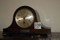 Front view of the wooden mantel clock showing the metallic clock face with black Arabic numerals and three black hands. The clock has a wooden case with a rounded top and decorative edge.