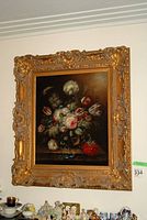 Framed oil painting hanging on wall above shelf with porcelain figurines and cups.