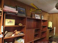 View of wooden shelves holding books, plaques, carved duck figurines, and a framed airplane inset. A model airplane sits on top of the shelving.