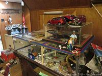 Wide shot of glass display case showing multiple model cars on top, fabric patches on base, figurines and large model airplanes inside