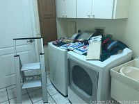 Laundry/utility room showing white aluminum step ladder near a white washer-dryer unit with mop, mop head, vacuum bags, chair covers and clothespins placed on top of dryer.