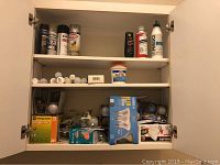Cabinet shelf containing multiple spray paint cans, adhesive containers and packages, and loose light bulbs in boxes and packs