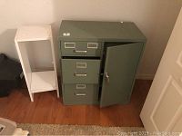 Photo showing green metal file cabinet with safe door open, next to white wooden shelf and electronic device
