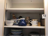 Shelf view showing a chicken-themed ceramic soup tureen with lid, a blue striped pitcher, white soup bowls, and decorative ceramic mugs and containers.