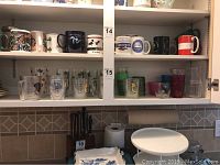 Various mugs and glasses arranged on shelves including ceramic mugs with designs, clear plastic Tervis tumblers with prints, and glassware.