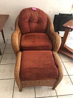 Rattan armchair with brown fabric cushions and matching ottoman in front, placed on tiled floor beside wooden furniture.
