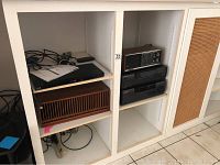 Photo showing shelving unit with Panasonic Blu-ray player on top shelf, vintage Panasonic wooden radio on the middle shelf, and Word multiplayer CD player below the radio.