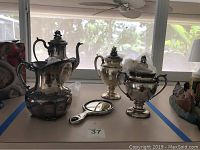 Overview of the silver plated urn, teapots, and mirror on a table near a window.
