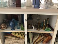 Two shelves with various decorative glassware and textiles. Blue and green vases, brass candlesticks, oil lanterns, folded textiles below.